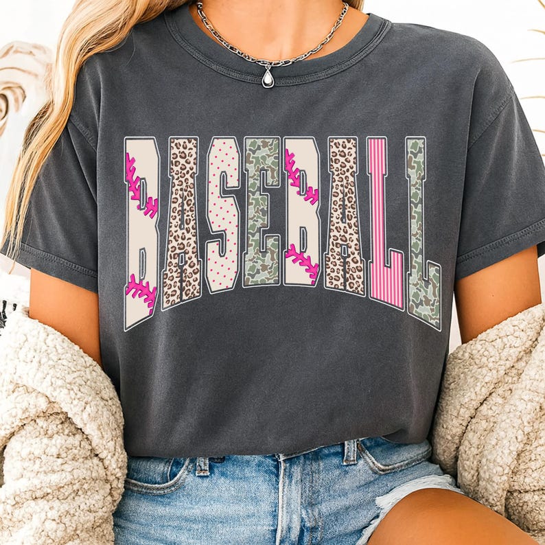 May include: Dark gray t-shirt with the word "BASEBALL" in a bold, decorative font. The letters feature baseball-themed patterns, including pink stitching, leopard print, and camouflage. The shirt has a crew neck and short sleeves.