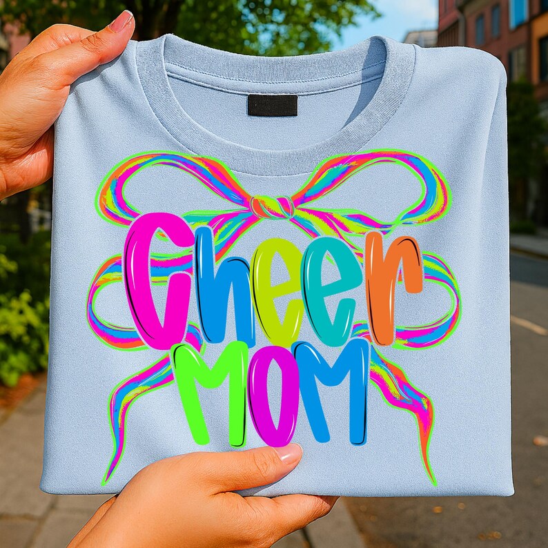 May include: A light blue crew neck t-shirt with the words "Cheer MOM" in colorful, 3D bubble letters. A rainbow-colored bow design surrounds the text. The shirt is held up by a person's hands.