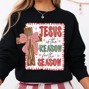 May include: Black sweatshirt featuring a decorative cross with a red and white plaid bow. The design includes the text "JESUS is the REASON for the SEASON" in red and green, with snowflake accents. A festive holiday-themed apparel item.