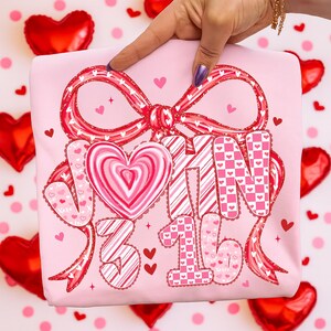 May include: A light pink t-shirt with a large red bow and the words "JOIN 3 16" in a decorative font. The letters and numbers are decorated with hearts and stripes. Small red hearts are scattered around the design.