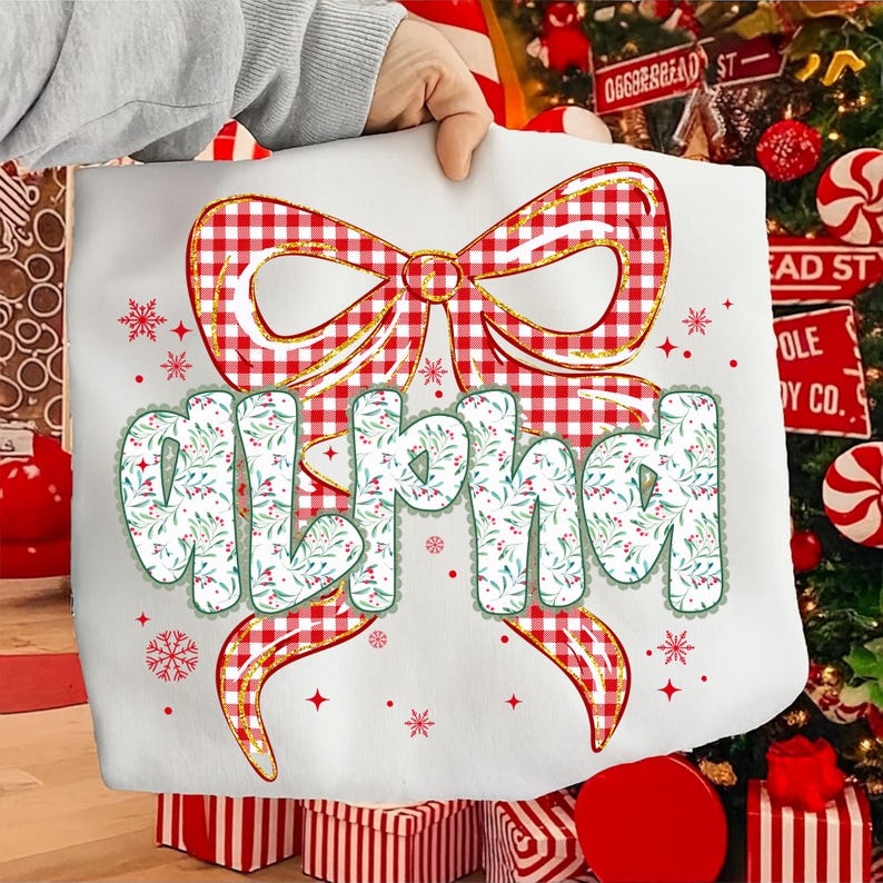 May include: White tote bag featuring a large red and white gingham bow with gold trim. The word "alpha" is printed in a whimsical font with a floral design. Snowflakes and stars are scattered around the design.