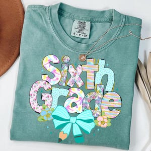 Op de afbeelding: Een turquoise Comfort Colors t-shirt met de woorden "Sixth Grade" in een kleurrijk, grillig ontwerp. De tekst bevat bloemen- en pastelpatronen, een turquoise strik en een potloodafbeelding. Een zilveren ketting is over het shirt gedrapeerd.