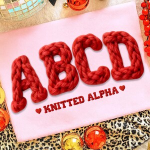 May include: A pink surface displays the letters ABCD in a chunky, knitted red yarn. Below the letters, the text "KNITTED ALPHA" is embroidered in red with a heart symbol. The background includes festive decorations.