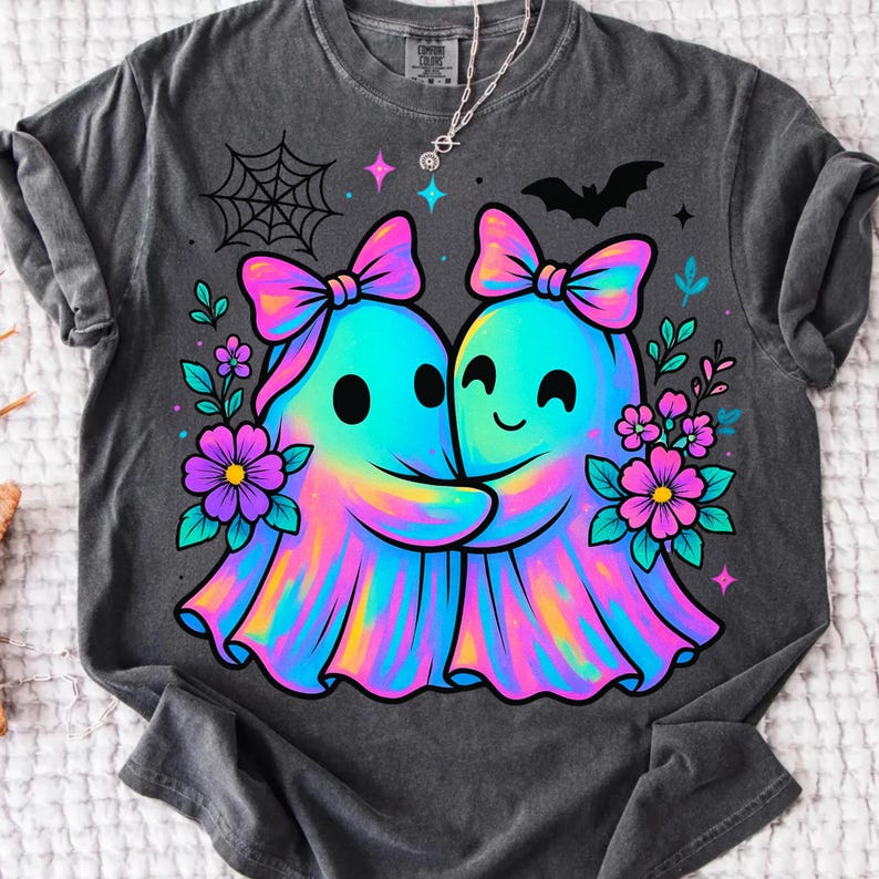 May include: Dark gray t-shirt featuring a colorful illustration of two hugging ghosts with bows, surrounded by flowers, a spiderweb, and a bat. The ghosts have a rainbow gradient effect. A silver necklace is visible.