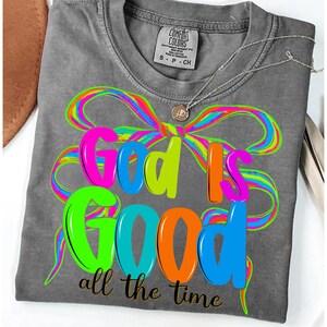 May include: A gray Comfort Colors t-shirt with the phrase "God Is Good all the time" in vibrant neon colors. The design includes a colorful bow and a silver necklace with a pendant.