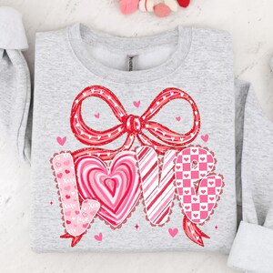 May include: A light grey sweatshirt featuring a large red bow and the word "LOVE" in a heart-shaped design. The letters are decorated with heart, stripe, and checkered patterns. Small pink hearts are scattered around the design.