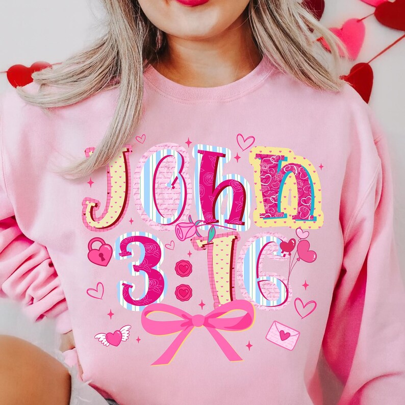 May include: Pink sweatshirt featuring the text "John 3:16" in a colourful, decorative font. The design includes hearts, a bow, and other Valentine's Day themed graphics. The sweatshirt is a solid pink colour.