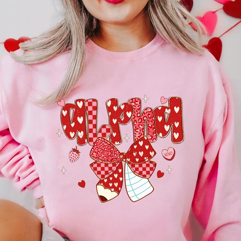 May include: Pink sweatshirt with the word "Alpha" in red, decorated with hearts, a bow, and a strawberry. The bow has a checkered pattern and a pencil design. The sweatshirt is a Valentine's Day theme.