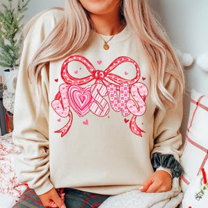May include: A cream-colored sweatshirt featuring a large graphic that says "JESUS" in pink letters. The letters are decorated with hearts and patterns, and the word is framed by a red bow and ribbons. Small pink hearts are scattered around the design.