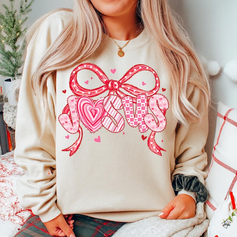 May include: A cream-colored sweatshirt featuring a large graphic that says "JESUS" in pink letters. The letters are decorated with hearts and patterns, and the word is framed by a red bow and ribbons. Small pink hearts are scattered around the design.
