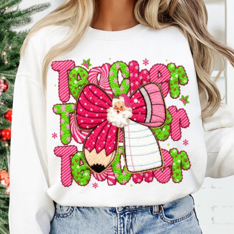 May include: White sweatshirt with a colorful graphic design. The design features the word "Teacher" in green and pink, a pink bow with a Santa Claus face, a pencil, and a notepad. The sweatshirt is a casual style.