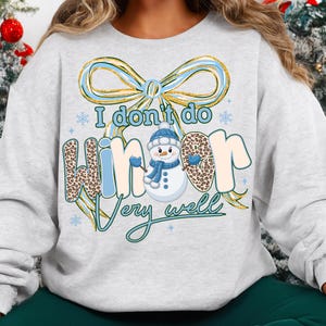 May include: A light gray sweatshirt with a winter-themed graphic. The design features a snowman wearing a blue hat and scarf, with the text "I don't do winter very well." The word "winter" has a leopard print pattern.