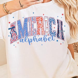 May include: White t-shirt featuring the word "AMERICA" in a patriotic design. The letters are filled with red, white, and blue patterns, including polka dots and floral prints. The word "alphabet" is written below in a matching blue font.