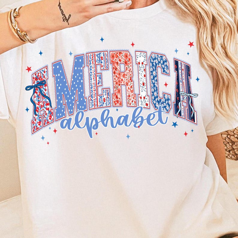 May include: White t-shirt featuring the word "AMERICA" in a patriotic design. The letters are filled with red, white, and blue patterns, including polka dots and floral prints. The word "alphabet" is written below in a matching blue font.