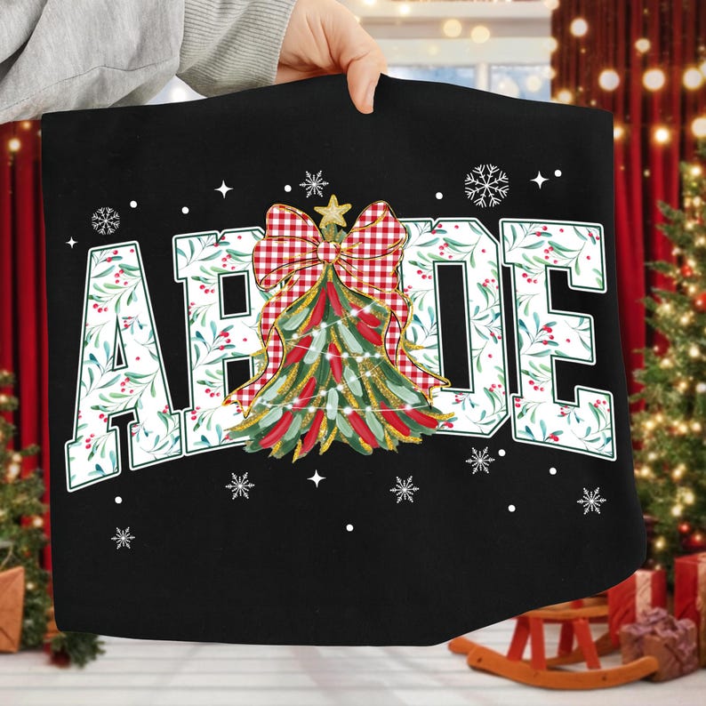 May include: Black Christmas-themed tote bag with the word "ABODE" in large white letters. The letters are decorated with green leaves and red berries. A Christmas tree with a red and white plaid bow is in the center, with snowflakes scattered around.