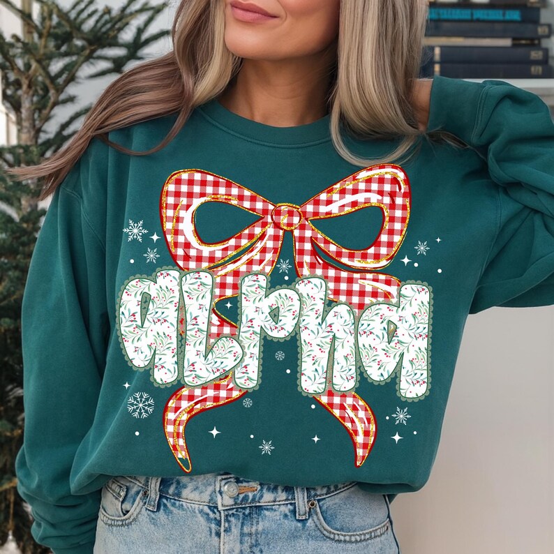 May include: A teal sweatshirt with a large red and white gingham bow design. The word "ALPHA" is printed in white, with a floral pattern and red berries. Snowflake and star accents complete the festive look.