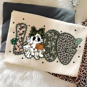May include: Cream-colored sweatshirt with the word "LOVE" in a mix of camouflage and leopard print. The letter "O" features a ghost holding a pumpkin and making a peace sign. Black stars and lace trim surround the letters.