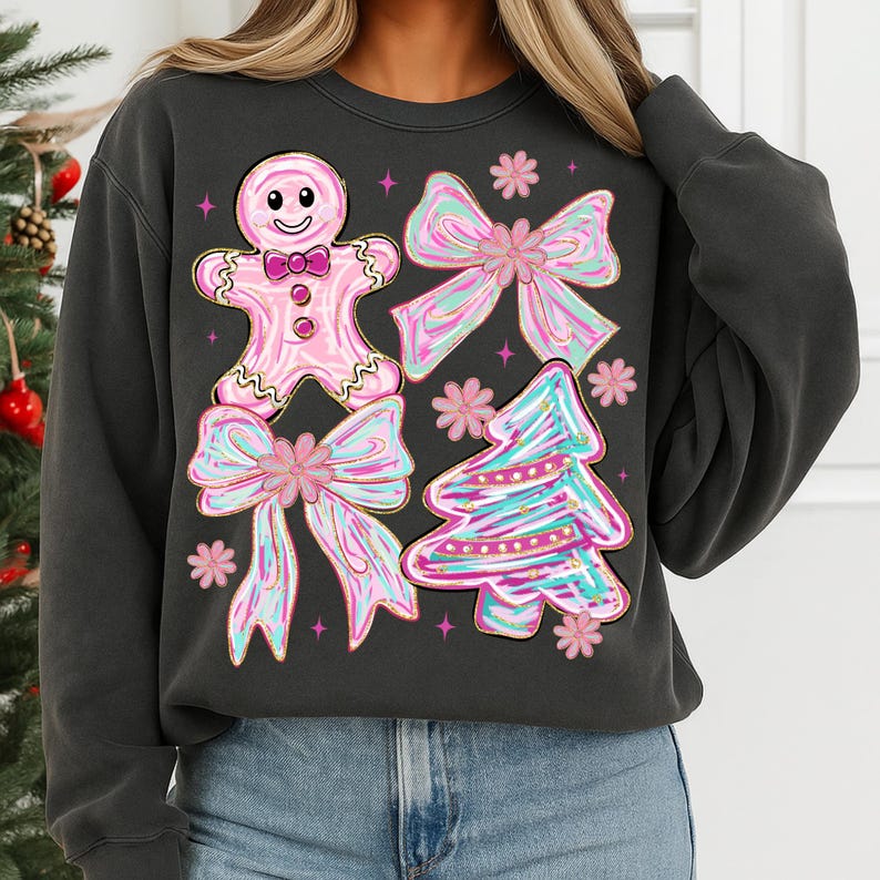 May include: Dark grey sweatshirt with a festive design of a gingerbread man, bows, and a Christmas tree in pink, blue, and gold. The design includes pink flowers and star accents, ideal for the holiday season.