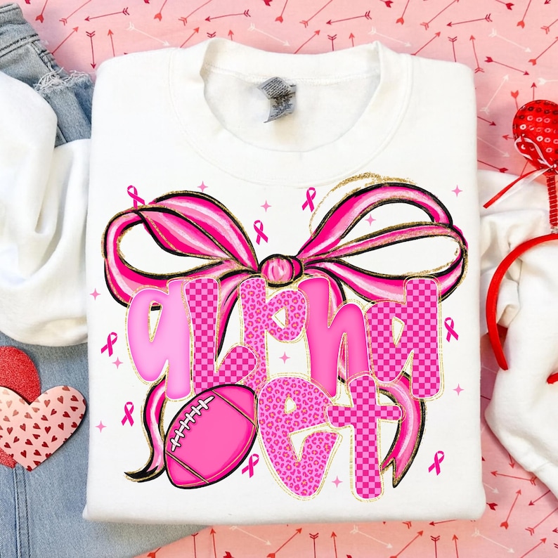 May include: White sweatshirt with a large pink bow and the words "Alpha Girl" in pink, with a pink football. The design includes pink ribbons and gold accents. A stylish and eye-catching design.
