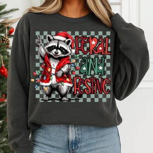 May include: Dark gray sweatshirt featuring a raccoon wearing a Santa hat and red vest, holding up a peace sign. The text "Feral and Festive" is displayed in red and green, with a checkered background. Christmas-themed apparel.