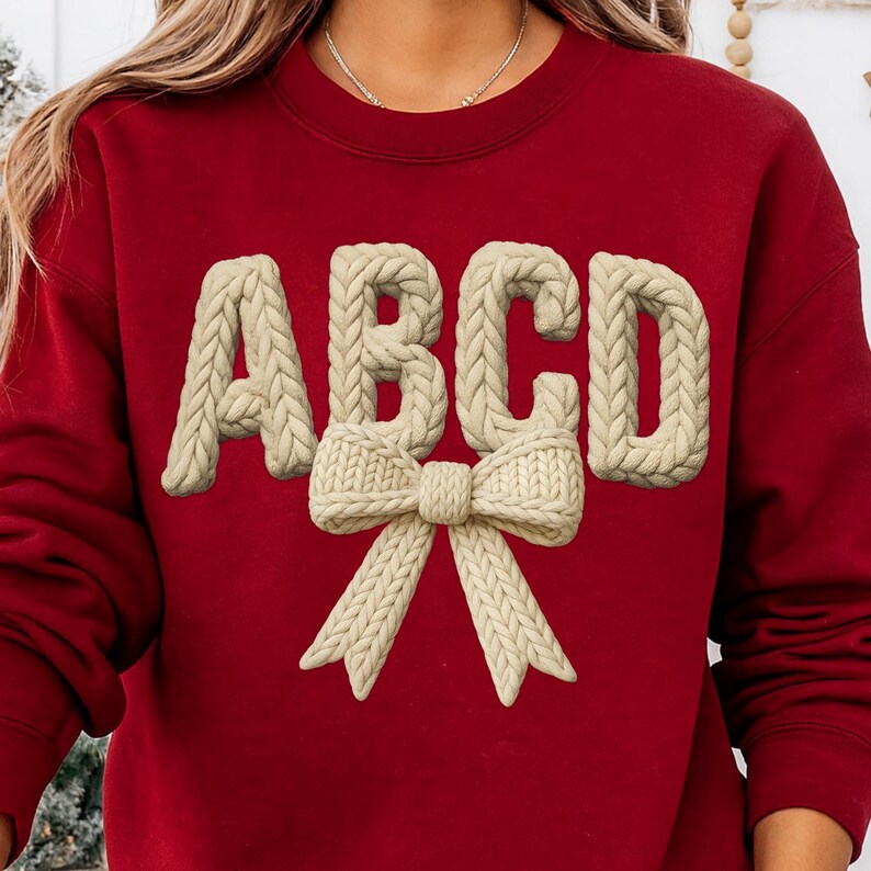 May include: A red sweatshirt featuring the letters ABCD in a cream-colored, knitted style. A cream-colored knitted bow is positioned below the letters. The sweatshirt is a solid, deep red color.