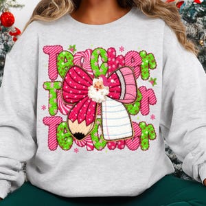 May include: A light gray sweatshirt with a festive design for teachers. The design features the word "Teacher" in green and pink, a pink bow with a Santa Claus, and a pencil. Small stars and snowflakes are scattered around the text.