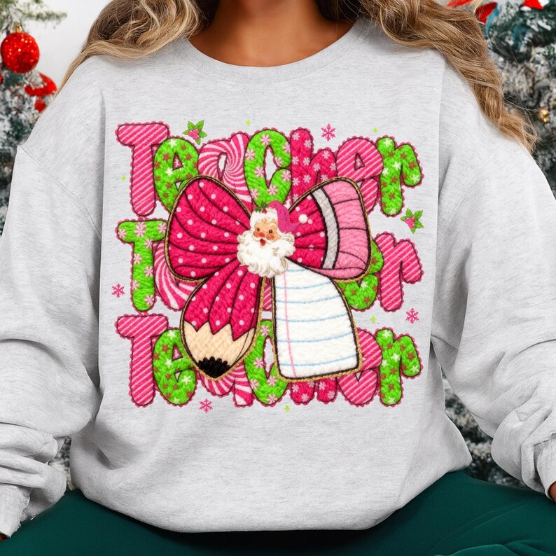 May include: A light gray sweatshirt with a festive design for teachers. The design features the word "Teacher" in green and pink, a pink bow with a Santa Claus, and a pencil. Small stars and snowflakes are scattered around the text.