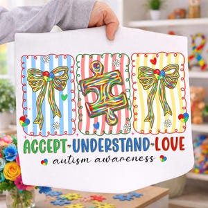以下が含まれることがあります： カラフルなイラストとテキストが描かれた白い布。デザインには2つのリボン、パズルのピース、そして「ACCEPT-UNDERSTAND-LOVE」の文字と、その下に「autism awareness」が含まれています。背景にはストライプと装飾的な要素があります。