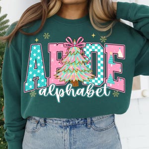 May include: Dark green sweatshirt with the word "AEIOE" in pink and teal, with a Christmas tree graphic. The word "alphabet" is written below in a cursive font. Snowflake and star graphics are also present.