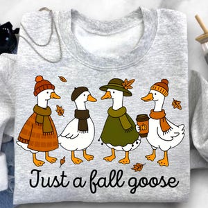 May include: A light gray sweatshirt featuring a whimsical illustration of four geese dressed in fall attire. The geese wear hats, scarves, and coats, with one holding a coffee cup. The text "Just a fall goose" is printed below the geese.