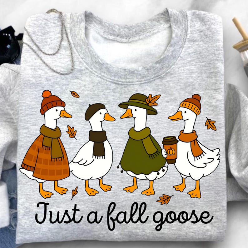 May include: A light gray sweatshirt featuring a whimsical illustration of four geese dressed in fall attire. The geese wear hats, scarves, and coats, with one holding a coffee cup. The text "Just a fall goose" is printed below the geese.