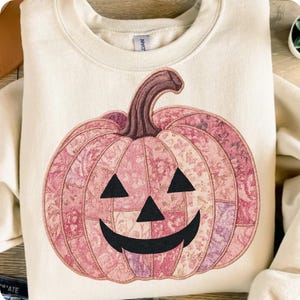 May include: Cream-colored sweatshirt featuring a pink pumpkin design. The pumpkin has a patchwork pattern with floral details, a brown stem, and a carved jack-o'-lantern face with black triangles for eyes and a curved mouth. A festive fall-themed garment.