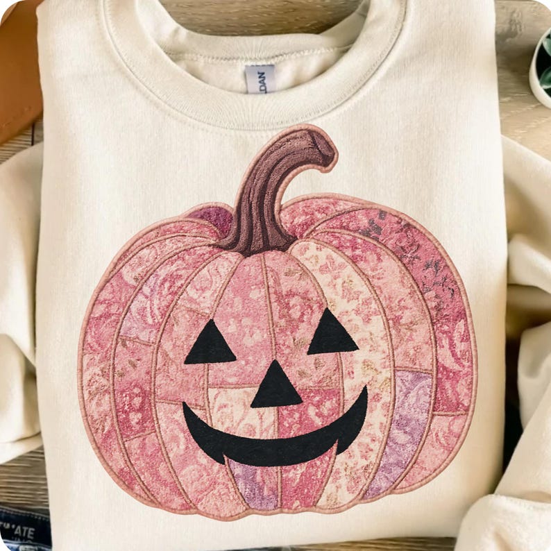K&ouml;nnte beinhalten: Cremefarbenes Sweatshirt mit einem rosa K&uuml;rbisdesign. Der K&uuml;rbis hat ein Patchwork-Muster mit floralen Details, einen braunen Stiel und ein geschnitztes K&uuml;rbisgesicht mit schwarzen Dreiecken f&uuml;r Augen und einem gebogenen Mund. Ein festliches Kleidungsst&uuml;ck zum Thema Herbst.