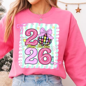 May include: Pink sweatshirt with a colorful graphic design. The design includes the numbers "26", a disco ball, champagne glass, and the text "Happy New Year". The design is outlined with a light blue and white striped border.