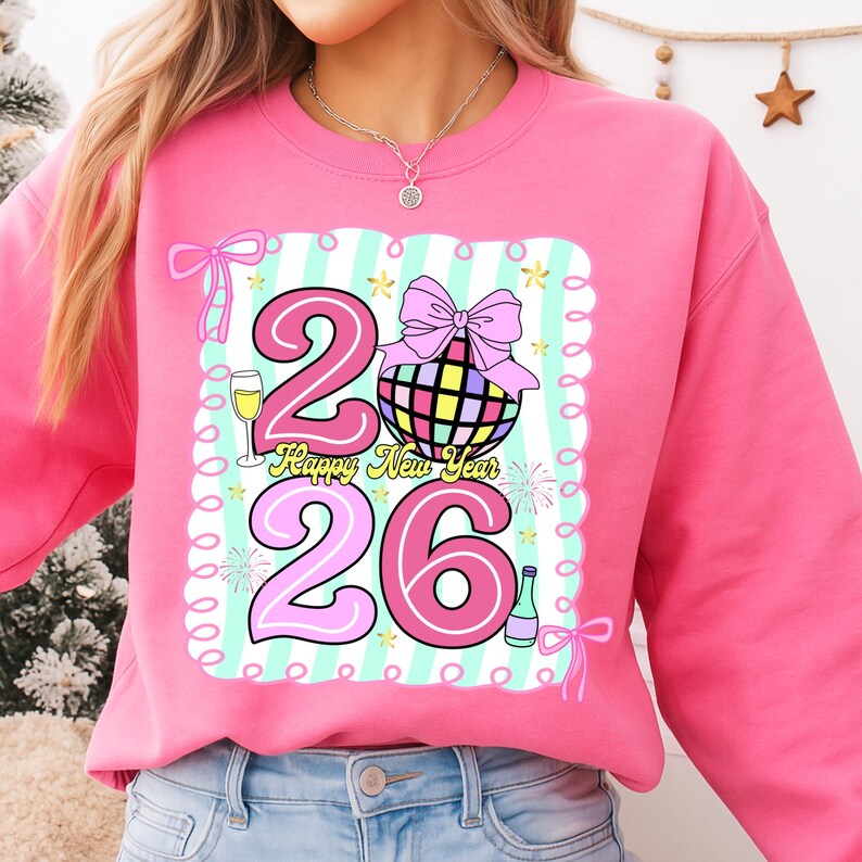 May include: Pink sweatshirt with a colorful graphic design. The design includes the numbers "26", a disco ball, champagne glass, and the text "Happy New Year". The design is outlined with a light blue and white striped border.