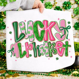 Pu&ograve; includere: Tessuto bianco con le parole "LUCKY ALPHABET" in verde e rosa. Le lettere presentano vari motivi, tra cui trifogli e strisce. Fiocchi verdi e stelle rosa decorano il design. Il tessuto &egrave; tenuto da una persona.