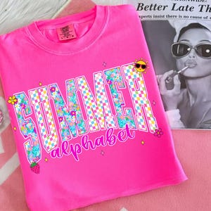 May include: A vibrant pink t-shirt with the words "SUMMER alphabet" in a floral and checkered design. The shirt features a cartoon sun, flowers, and a strawberry graphic. The shirt is made of a soft material.