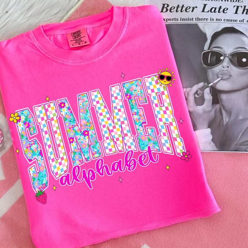May include: A vibrant pink t-shirt with the words "SUMMER alphabet" in a floral and checkered design. The shirt features a cartoon sun, flowers, and a strawberry graphic. The shirt is made of a soft material.