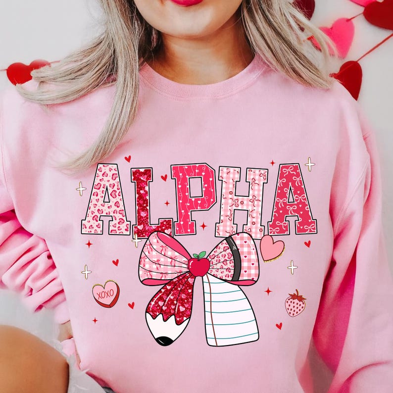 Op de afbeelding: Roze sweatshirt met het woord "ALPHA" in een mix van patronen, waaronder luipaardprint en strikken. Een grote strik met een potlood- en notitieboekontwerp is onder het woord gecentreerd. Kleine hartjes en een aardbei zijn verspreid.