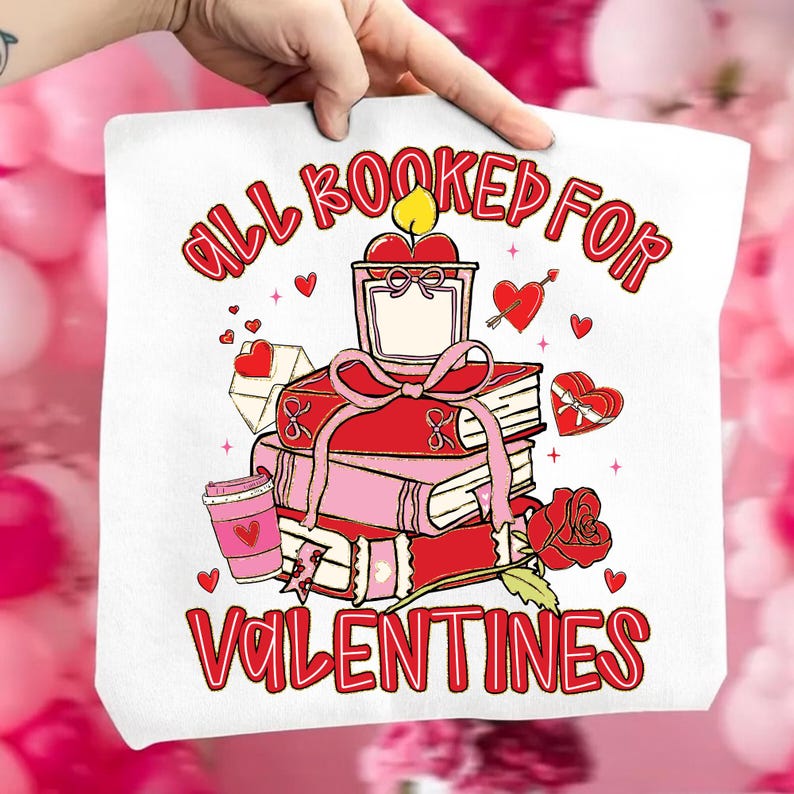 May include: White tote bag with a Valentine's Day design. The design includes a stack of books, a candle, a rose, and the text "ALL BOOKED FOR VALENTINES" in red. Pink hearts and a coffee cup are also featured.