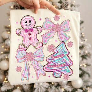 May include: Cream-coloured t-shirt with a gingerbread man, bows, a Christmas tree, and floral accents. The designs are in pink, teal, and gold. The gingerbread man has a smiling face and bow tie. The bows and tree have a watercolour effect.