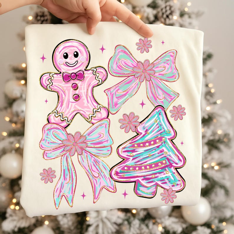 May include: Cream-coloured t-shirt with a gingerbread man, bows, a Christmas tree, and floral accents. The designs are in pink, teal, and gold. The gingerbread man has a smiling face and bow tie. The bows and tree have a watercolour effect.