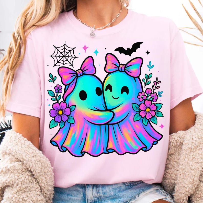 May include: Pink t-shirt featuring a colorful graphic of two hugging ghosts with bows, surrounded by flowers, a spiderweb, and a bat. The ghosts have a rainbow tie-dye effect. A cute and whimsical design.