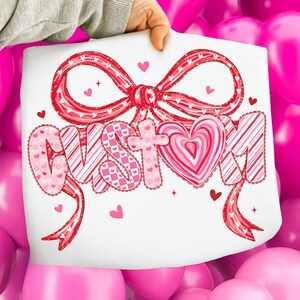 May include: A white sheet with the word "CUSTOM" in pink, red, and white, with a large bow and heart accents. The letters have various patterns, including hearts and stripes. Small hearts and stars are scattered around the text.