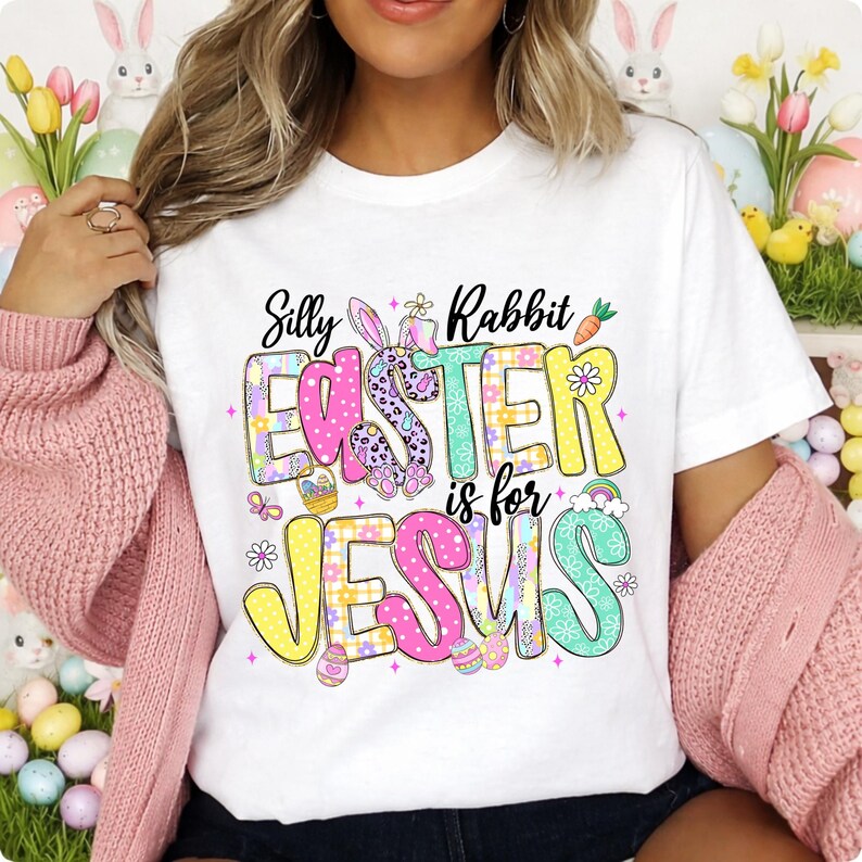 May include: White t-shirt with a colorful graphic that reads "Silly Rabbit Easter is for Jesus." The design features a bunny, Easter eggs, and flowers in pastel colors. The shirt is a casual, everyday wear item.