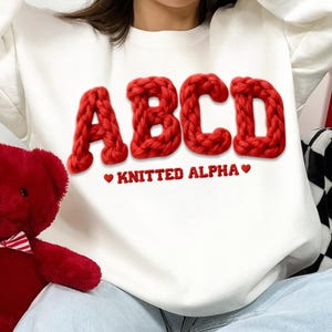 May include: White sweatshirt with large, red, knitted letters spelling "ABCD". Below the letters, the text reads "KNITTED ALPHA" in red. A red teddy bear is visible on the left side of the image.