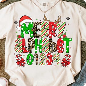 以下が含まれることがあります： クリーム色のTシャツに、お祝いのクリスマスのデザイン。グラフィックは、「MERRY ALPHABET 01234」の文字を、ヒョウ柄、キャンディケーンのストライプ、サンタ帽など、さまざまなスタイルで表現しています。雪の結晶とトナカイの角も含まれています。
