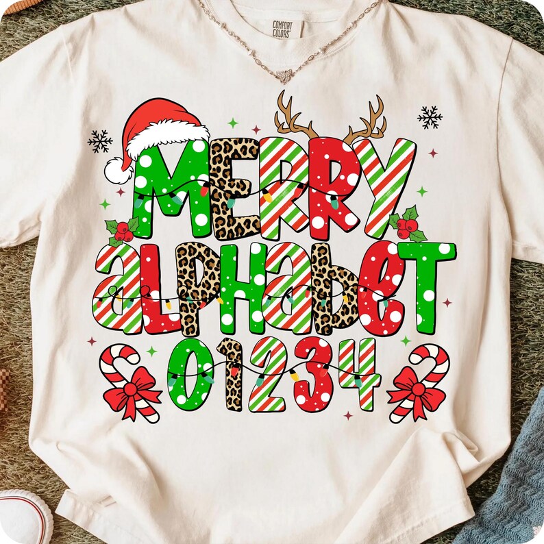 以下が含まれることがあります： クリーム色のTシャツに、お祝いのクリスマスのデザイン。グラフィックは、「MERRY ALPHABET 01234」の文字を、ヒョウ柄、キャンディケーンのストライプ、サンタ帽など、さまざまなスタイルで表現しています。雪の結晶とトナカイの角も含まれています。