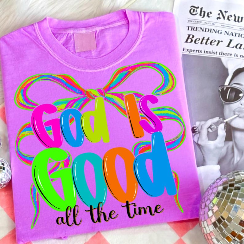 May include: A lilac-colored t-shirt with the phrase "God is Good all the time" in vibrant, multi-colored bubble letters. A rainbow-colored bow design arches over the words. The shirt is a casual wear item.