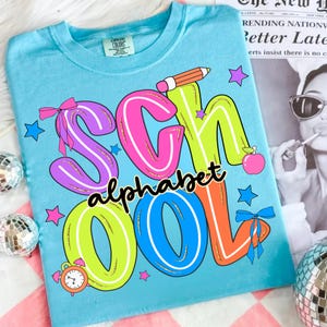 May include: A light blue t-shirt with a colourful graphic that reads "SCHOOL alphabet COOL". The word "SCHOOL" is in large, outlined letters with bows and stars. A pencil, apple, and alarm clock are also featured.